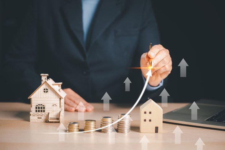 Businessman drawing rising arrow over coins and house models, representing real estate investment growth.
